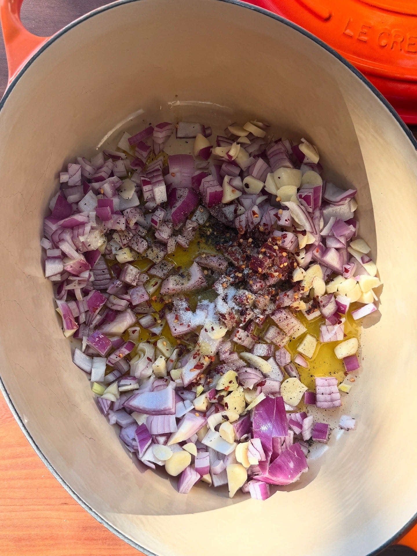 Chopped red onion, garlic, olive oil, salt, pepper, and red pepper flakes in a cream-colored pot, ready for cooking.