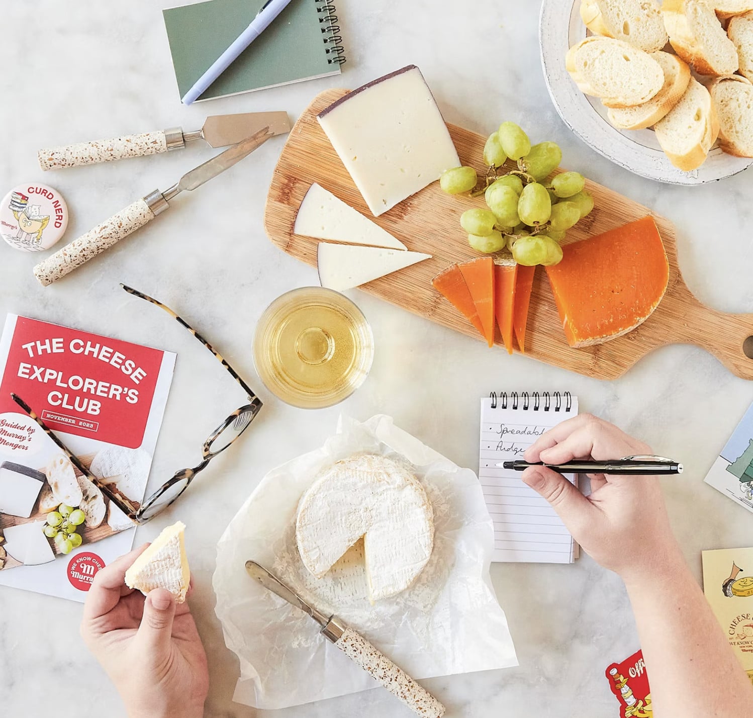 A person writes notes at a table with various cheeses, a round of brie, sliced bread, green grapes, a glass of white wine, and notebooks on a marble surface—a cozy scene inspired by GrossyPelosi.