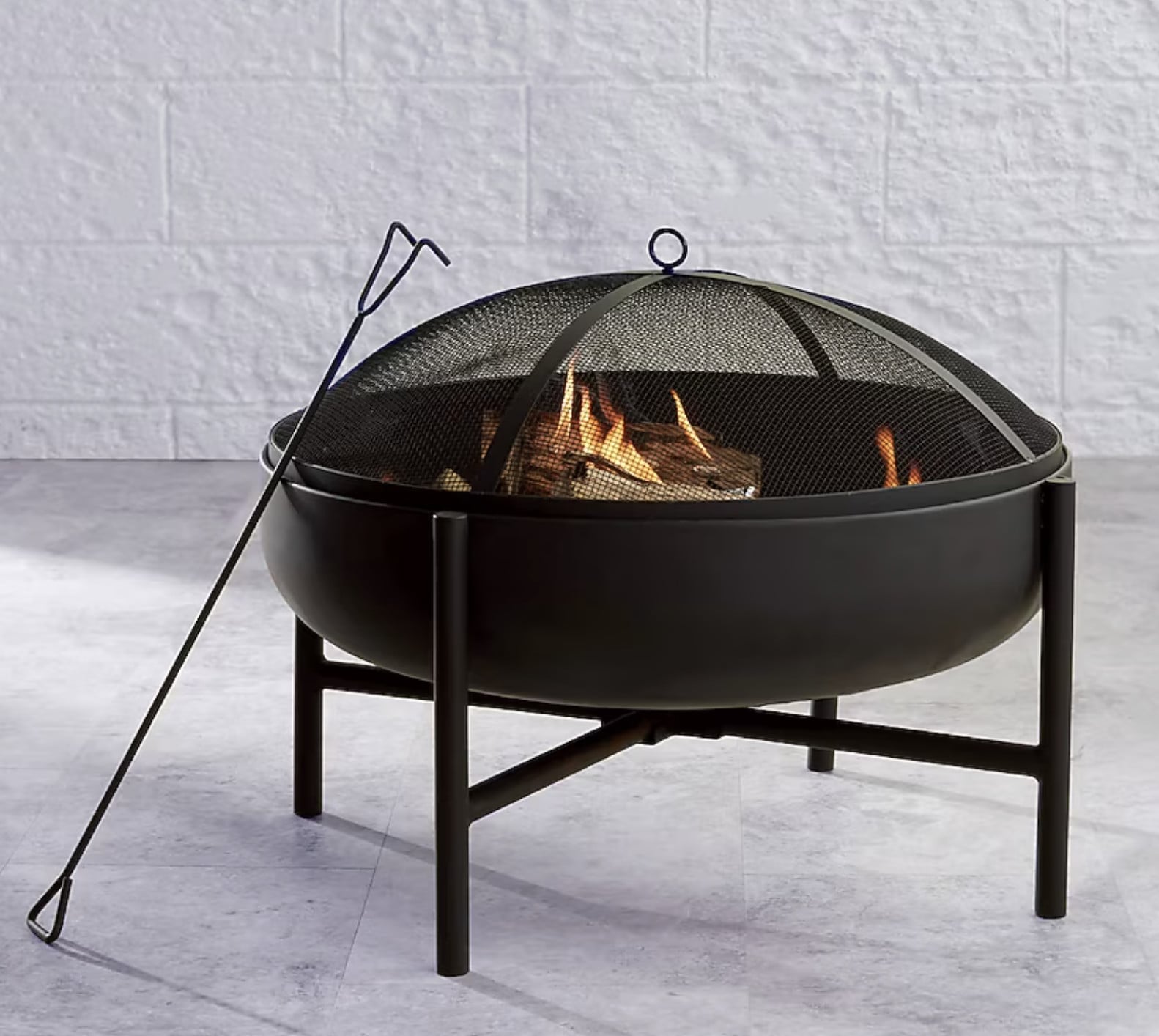 A black metal outdoor fire pit with a mesh screen cover and a metal poker tool beside it, containing burning wood and flames.