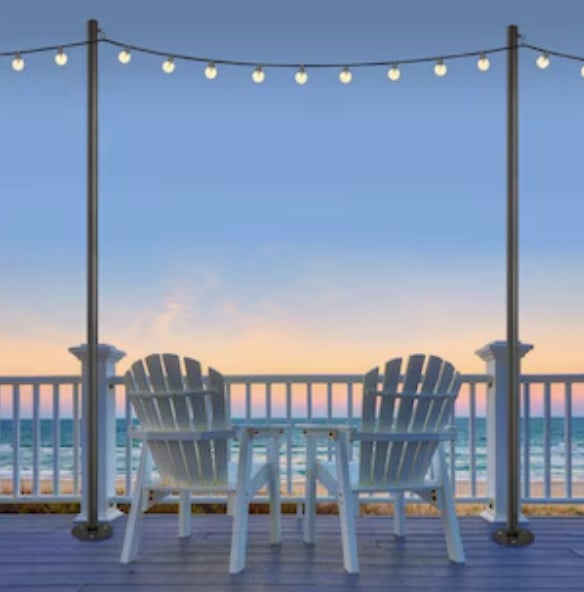 Two white Adirondack chairs and a small table on a deck facing the ocean at sunset, with string lights hanging above.