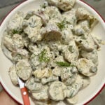 A bowl of creamy potato salad with herbs and capers, garnished with dill, served with a red-handled fork.