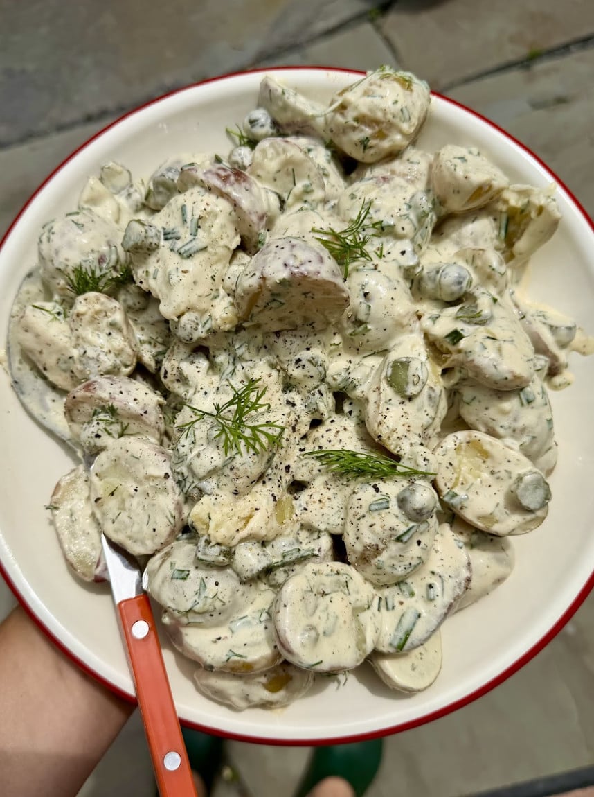 A bowl of creamy potato salad with herbs and capers, garnished with dill, served with a red-handled fork.