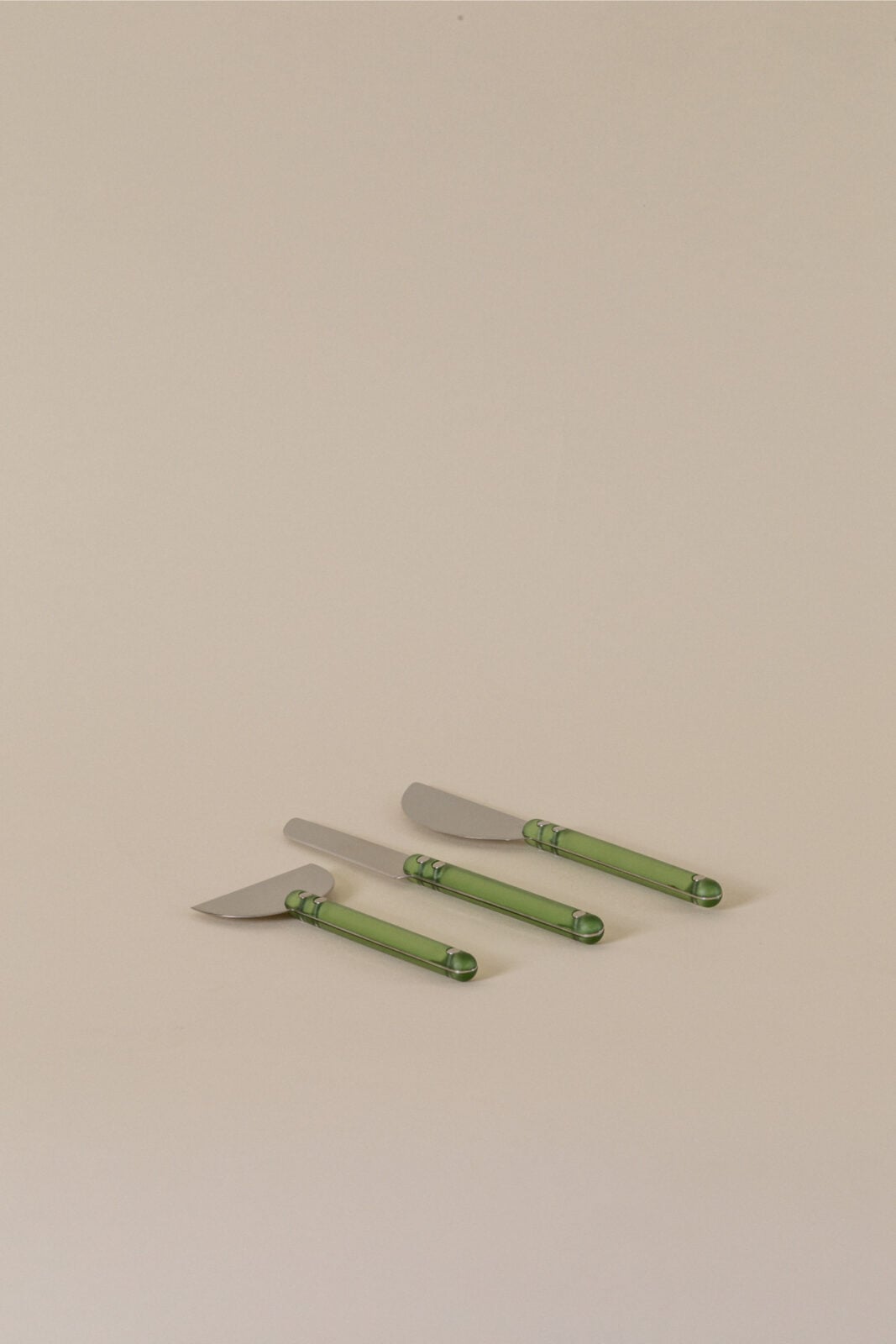 Three butter knives with green handles, inspired by Dan Pelosi’s signature GrossyPelosi style, are arranged in a row on a neutral beige surface.