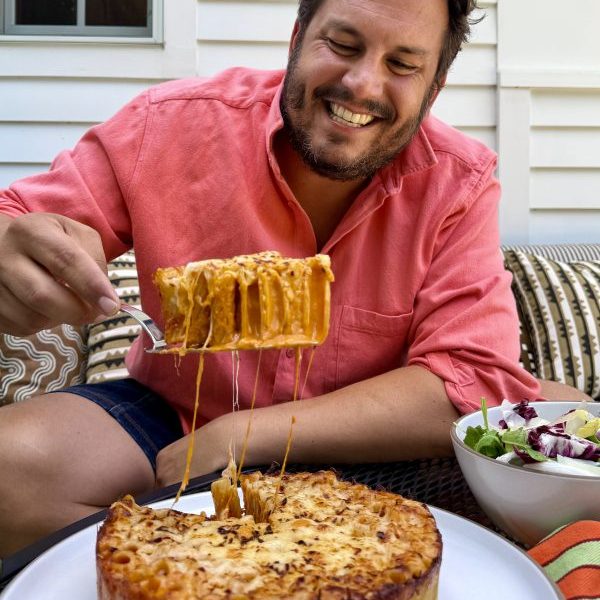 A man in a pink shirt smiles while lifting a cheesy pasta slice from a round baked pasta dish, with a salad bowl on the table.