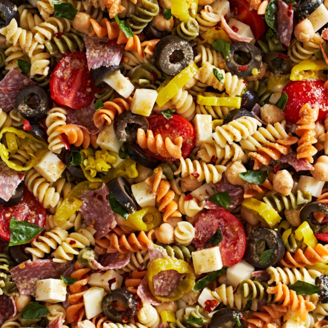 Close-up of colorful pasta salad with rotini noodles, cherry tomatoes, salami slices, black olives, cheese cubes, chickpeas, peppers, and chopped greens.