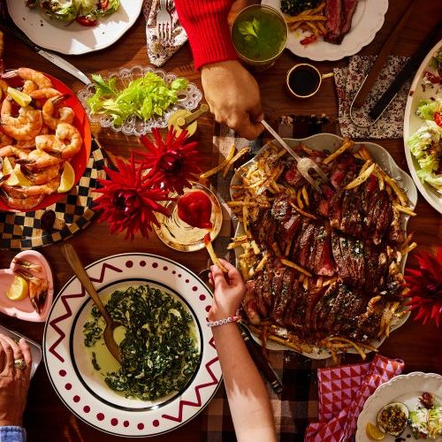 A table set with various dishes including shrimp, steak with fries, deviled eggs, spinach, salad, and drinks, with several people serving and eating food.