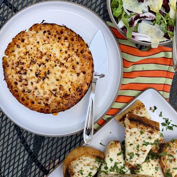 A round baked pasta dish on a plate with a serving spatula, next to a green salad and garlic bread slices on a metal outdoor table.