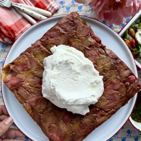 A square rhubarb cake topped with a large dollop of whipped cream on a white plate, with a hand holding the plate over a patterned tablecloth.