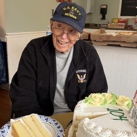 An elderly man wearing a U.S. Navy Veteran cap sits at a table with a birthday cake labeled "104" and a slice of cake on a plate in front of him.