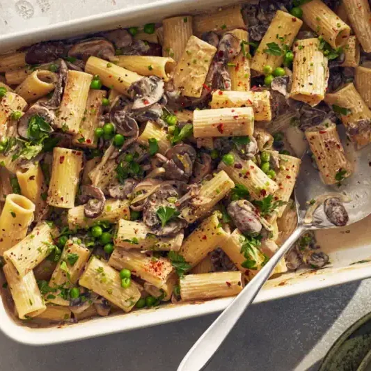 Rigatoni pasta with mushrooms and peas in a creamy sauce, topped with herbs and red pepper flakes, served in a white baking dish with a serving spoon.