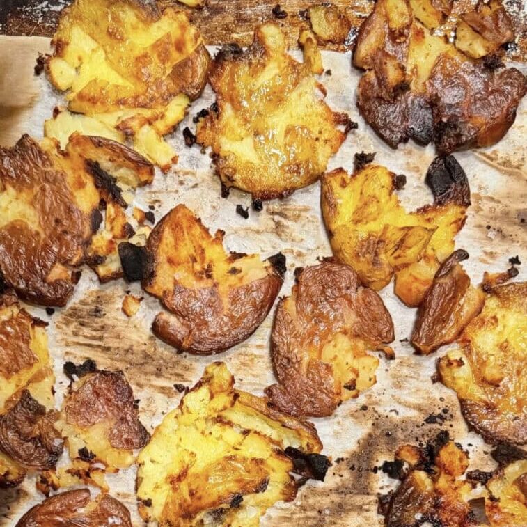 Crispy smashed potatoes with golden brown edges and charred spots on a parchment-lined baking tray.