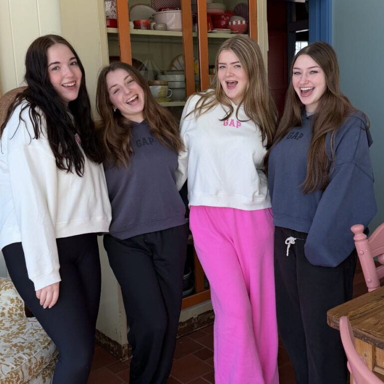 Four young women stand indoors, smiling at the camera. They wear GAP sweatshirts and casual pants, with a glass-fronted cabinet and pink chair visible in the background.