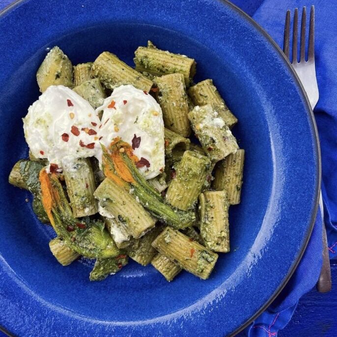 Rigatoni pasta with green pesto, zucchini blossoms, and two dollops of ricotta, garnished with red pepper flakes, served on a blue plate with a fork and napkin.