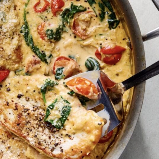 A skillet containing creamy chicken breasts cooked with spinach and cherry tomatoes, with a spatula lifting one piece.