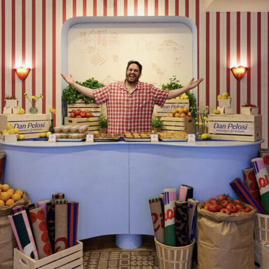 A person in a red-checkered shirt stands smiling with arms outstretched behind a counter with fresh produce, crates, and colorful rolled mats in a shop with red-striped walls.
