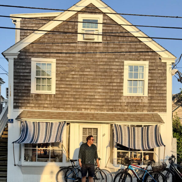 A person stands in front of a shingled two-story building with striped awnings and several bicycles parked outside, under clear sky and sunlight.