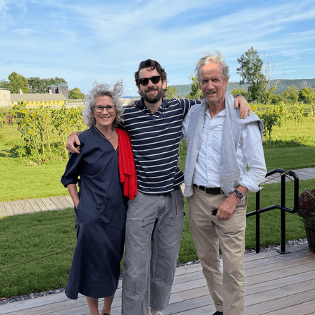 Three people stand side by side outdoors on a wooden deck, smiling at the camera with greenery and a blue sky in the background.