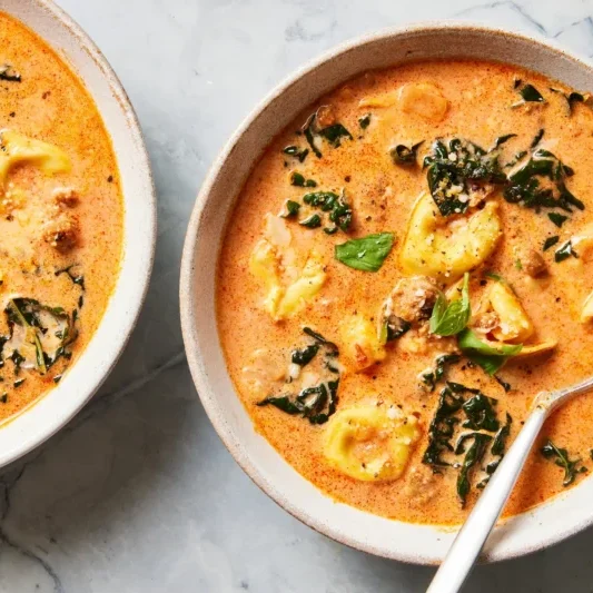 Two bowls of creamy tomato-based soup with tortellini, sausage, and spinach, garnished with fresh basil, sit on a marble surface. A spoon rests in one of the bowls.