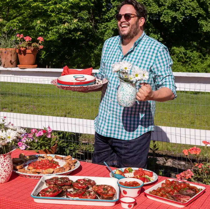 A man in a checkered shirt stands outdoors by a table with various dishes and flowers, holding a plate and a jug of daisies, smiling with greenery in the background.